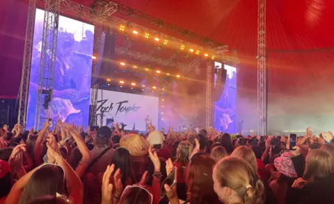 A crowd watches Zach Templar play on stage in a large red big top-style tent. He can be seen playing a guitar on screens either side of the stage