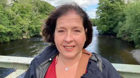 Karen standing on a bridge above a river. She is positioned in the centre of the frame and is facing the camera. She has dark, shoulder-length hair and is wearing a red V-neck top underneath a dark waterproof jacket. A brown cross-body strap runs diagonally across the front of the jacket, and a small silver necklace with a pendant is visible around the neck.