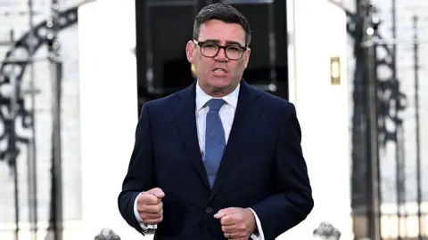Andy Burnham speaks outside 10 Downing Street after becoming prime minister. He is wearing black glasses, a navy blue suit, white shirt, and blue cotton tie.