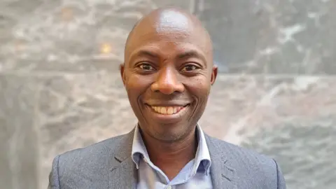 head and shoulders of man, looking astatine  camera, bald and cleanable  shaven, smiling, wearing grey suit