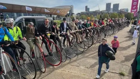 A long row of riders sat on Penny Farthing bicycles hold each other's handle bars. 