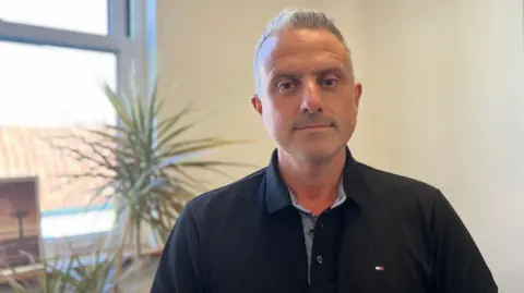 A man with grey hair and a black polo shirt, with the top button undone, is looking into the camera with a straight face. The sun is shining through a window behind him and there is a pot plant to the left of the photo beside the window, and a framed picture on the window sill