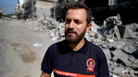 Goktay Koraltan Hussein Jaber spoke to the camera. He is a slim man wearing a blue polo shirt, standing on the street. Gray debris from a damaged building littered the road behind him.