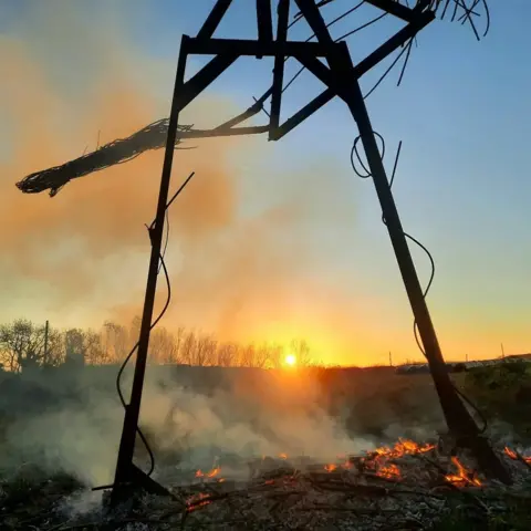 Trevor Leat A metal frame with some wicker hanging from it, standing above a bed of smouldering ash at sunset