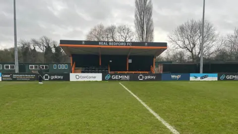 Tony Fisher/BBC A small football stand with Real Bedford FC written on the top. There are advertising hoardings surrounding it. The football pitch is in front of it and there two tall lamps either side of it.