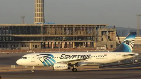 AirTeamImages A picture of a white and blue EgyptAir plane, stationary on a runway with concrete buildings behind it.