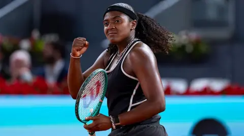 Hailey Baptiste, wearing a black vest and shorts, black headband and a ponytail, holds her green Wilson racket in her left hand and punches her right fist in celebration