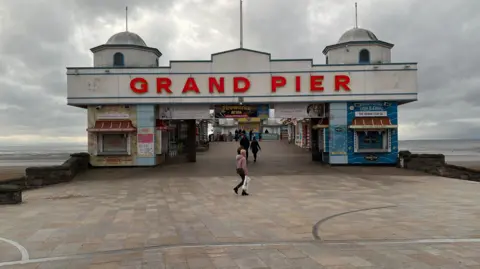 A beach pier which has the words 'Grand Pier' written on the front in red writing. A few people can be seen walking through the pier's entrance.