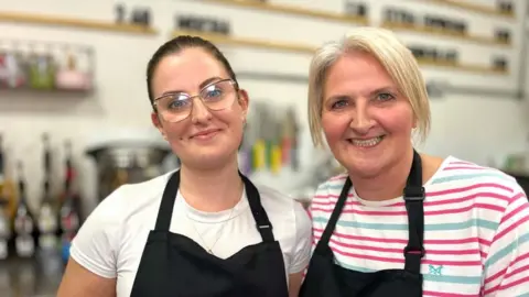 Martin Giles/BBC Mia and Sue Wallace are standing close to each other behind the counter of a coffee shop. There is a large menu on the wall in the background. Mia is wearing a white T-shirt and black apron. Sue is wearing a white, pink and green striped T-shirt with a black apron. 
