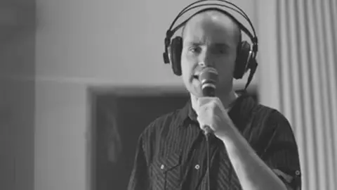 A man with a bald head, wears a headphone set and sings into the microphone in a studio 