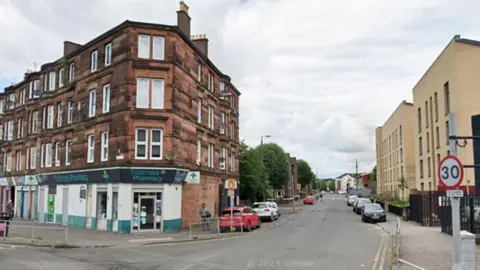 A junction on a city street, with rows of redbrick flats down one side of the street, above a pharmacy 