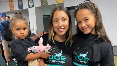 A woman with brown hair holds up a small girl in one arm while standing next to an older girl, both her daughters. They are smiling while standing inside what appears to be a club house. Each is wearing a black 'Joe Thompson Foundation' t-shirt. 