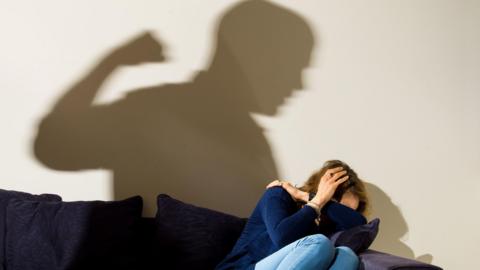 shadow of a man with a clenched fist as a woman covers in the corner. 