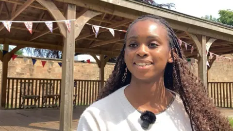 A girl with long braided black hair, wearing a white jumper, is standing in front of a wooden pavilion with Union Jack bunting around the outside.
