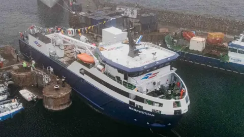Isles of Scilly Steamship Group An aerial view shows the Menawethan freight ship docked in a harbour beside smaller boats and quayside buildings, with cranes, equipment and workers visible around the busy port.