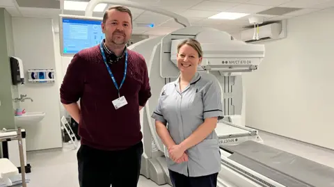 Lee Evans is Radiology Risk Manager at the Queen Elizabeth Hospital pictured with Hannah St Barbe Oldham, Nuclear Medicine Lead Radiographer. Lee is wearing a crimson knit jumper and black trousers. Hannah wears light grey scrubs and black trousers. 