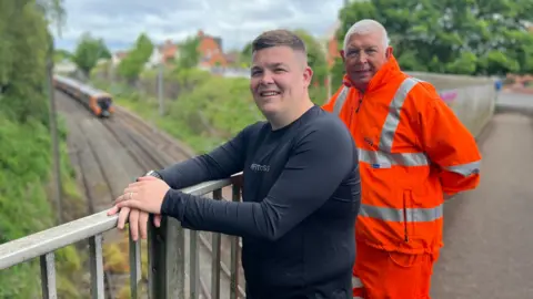 Two men stand on a railway bridge smiling at the camera. The man on the right wears an orange hi-vis National Rail jacket while the man on the left wears a black long-sleeved shirt and jeans. Below on the railway track, a train is on the line.