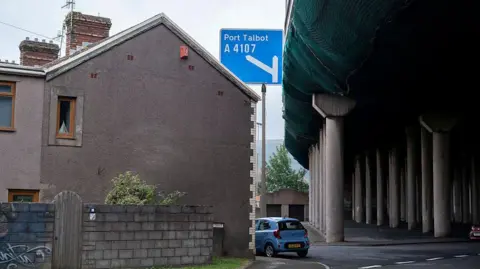 Getty Images The images shows the side of a grey end-of-terrace house behind a breeze block wall with a blue car parked outside and next to the pillars that hold up the flyover of the M4 motorway at Port Talbot