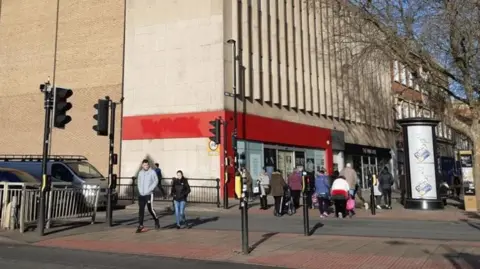 Local Democracy reporter Roadside view of the TK maxx building -with a red entrance to a large cream building. People can bee seen walking in front of the building onto the road.