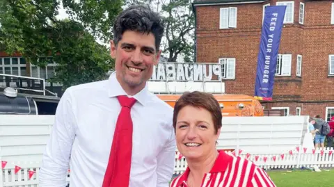 JACKIE STORER A woman and a man (former England cricket captain Alastair Cook) stand on a cricket pitch. The man is dressed in a white shirt and red tie. The woman is dressed in a red and white blouse. There is a food truck, trees and a house over the walled boundary behind them.