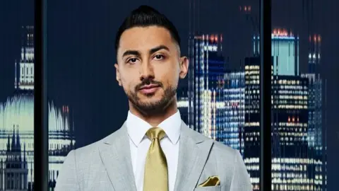 Rajan Gill is seen in a publicity photograph from The Apprentice. He is wearing a light grey suit with a white shirt and gold tie and a gold pocket square. He is in front of the window with the City of London and St Paul's Cathedral lit up at night behind him. The view of the city is slightly blurred in the glass.