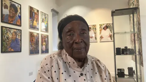 A woman sitting in an art gallery, with portrait photographs and vintage cameras on display in the background.
