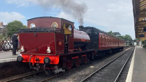 Jenny Kirk/BBC A red and black locomotive and carriages are stood still at a railway station. On the right is a platform and a bridge is in the background.