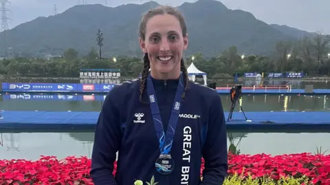 Beth Littlewood A woman with brown hair in two french plaits. She is wearing a blue sports jacket and has a medal around her neck. She is smiling and looking at the camera and is stood in front of a body of water with mountains in the background.