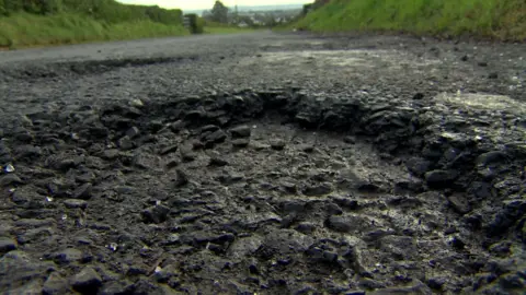 A hole in a dark gravel road up close with a road continuing in the background surrounded by fields