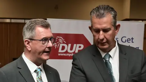 Jeffrey Donaldson and Edwin Poots pictured side by side. Both wearing grey suits. DUP poster in background.