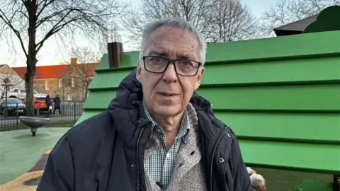 The photo shows a man with grey hair, wearing glasses and a black hooded coat standing in front of children's play equipment in a park. 