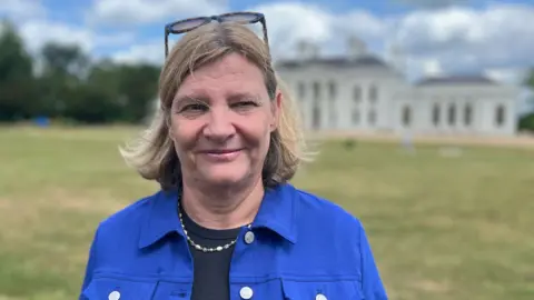 Qays Najm/BBC Councillor Natacha Dudley from Chelmsford City Council. She wears a blue jacket with navy blue t-shirt underneath. She has a white and blue necklace around her neck, and has sunglasses pushed upwards onto the top of her head. She has blonde shoulder-length hair. Cllr Dudley is stood in front of Hylands House in Hylands Park, Chelmsford.