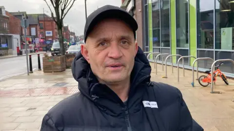 Dale Owusu poses for pictures on a street of shops in Levenshulme outside the sports centre. He wears a dark baseball cap and coat