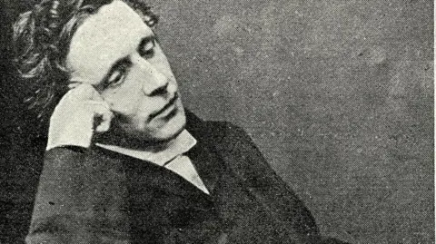Getty Images Archive photo of Lewis Carroll seated in a relaxed pose, wearing formal Victorian-era clothing including a dark suit and white shirt. He is resting one arm on the chair and the other near the face. 