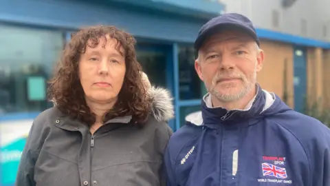 Ancel Mitchell, a middle-aged woman wearing a grey winter coat with brown curly hair standing next to Simon Elmore, a middle-aged an wearing a blue tracksuit top and a navy blue cap with a shirt grey beard. 