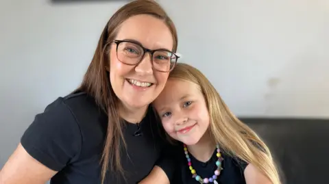 Mum Sara is smiling and wearing glasses. She is alongside her young daughter Ellie who is smiling and wearing a multi-coloured necklace. They are pictured at home when Ellie is educated by her mum. 