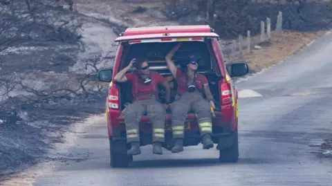 Two fire officers, one female, are in the back of a red fire vehicle with the boot door open. They are wearing fire safety clothing and around their necks are breathing masks. Both wear sunglasses. Around them are the remnants of burnt trees and grassland. The red vehicle appears to be driving on a road away from the camera.