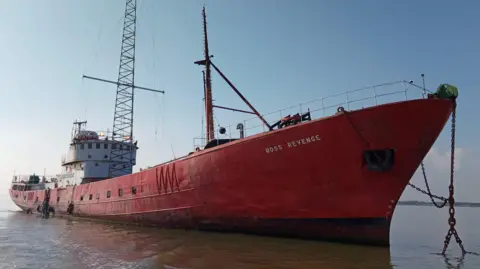 Owen Ward/BBC Ross Revenge, a bright red ship with a large aerial, lies at anchor.