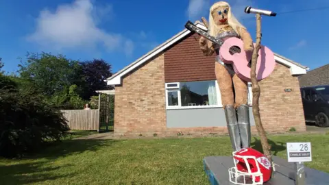 BBC Weather Watchers/Sibley A scarecrow holding a pink guitar with a red American football helmet in front.
