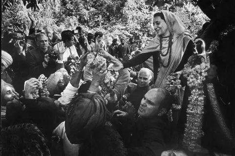 Raghu Rai/The India Today Group via Getty Images Indira Gandhi, wearing a saree and a beaded garland, in a crowd of people during an election campaign in 1977.
