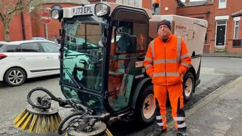 Man in his 60s stands on the pavement next to a clear-sided road sweeping vehicle with large yellow bristles on the front. He is wearing an orange high-visibility jacket and trousers and a black woolly hat.