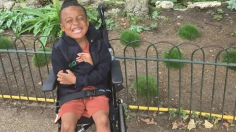 supplied A young boy in shorts and a black hoodie smiles as he sits in a wheelchair