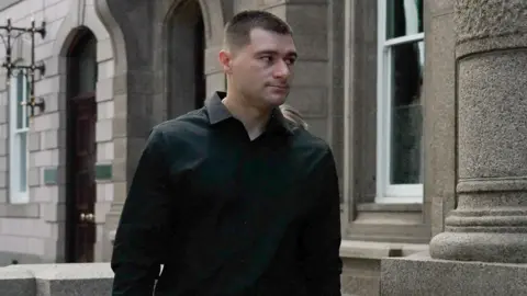 BBC Artur Sevash-Zade walks into Jersey Royal Court. He is wearing a black shirt. He has short black hair.