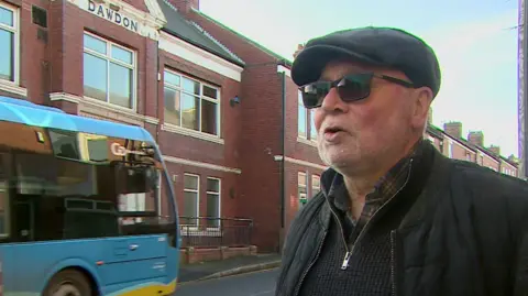 A grab taken from a TV package of Bob Arthur, an older man wearing a black flat cap, sunglasses and a black jacket. He is standing in front of the hall, a red brick building, and a bus is in the background.