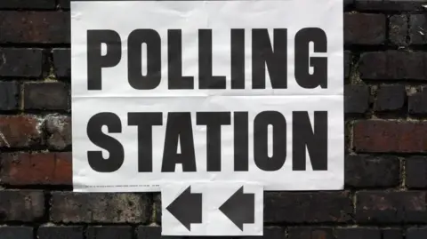 A white polling station sign with black letters and arrows pointing to the left. The sign is on a brick wall.