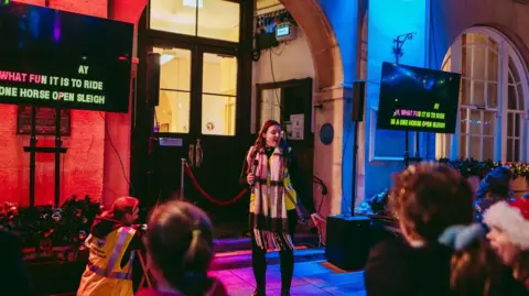 Kyle Stevens A woman stands in between two TV screens singing into a microphone. The screens have 'Oh What Fun It Is To Ride On A One Horse Open Sleigh' written on them. People are watching her sing and the back of their heads are in the bottom of the shot, one person has on a Christmas hat.