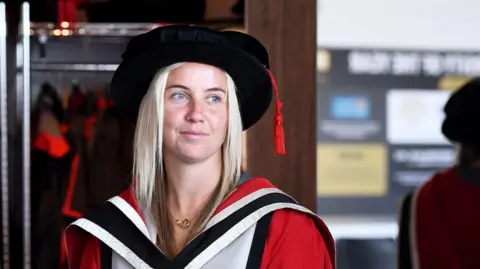 Beth Mead is wearing a black graduation cap and a red, black and white graduation gown. She has long blonde and brown hair and is looking off into the distance with a slight smile on her face. She is standing in front of a row of gowns and a mirror which is showing her reflection from behind.