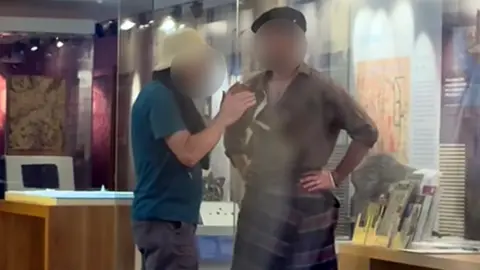 A man in a kilt with hands on hips and a man in a hat standing beside each other in a museum room, both have their faces blurred to hid their identities