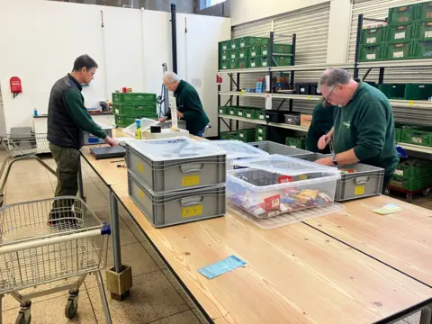 Amy Holmes/BBC A picture of three men packing food into crates in a warehouse in Dunstable. They are wearing green tops.