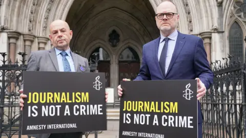 Barry on the left is bald, wearing a grey suit, blue shirt and blue tie. Trevor on the right is taller, has a bit more hair than Barry and is wearing black round glasses. He is wearing a blue shirt and dark blue navy tie under a dark navy suit. They are both holding black signs that read 'Journalism is not a crime, Amnesty International' on them. Behind them, is the Royal Courts of Justice entrance, a grand, pillar shape with big windows.